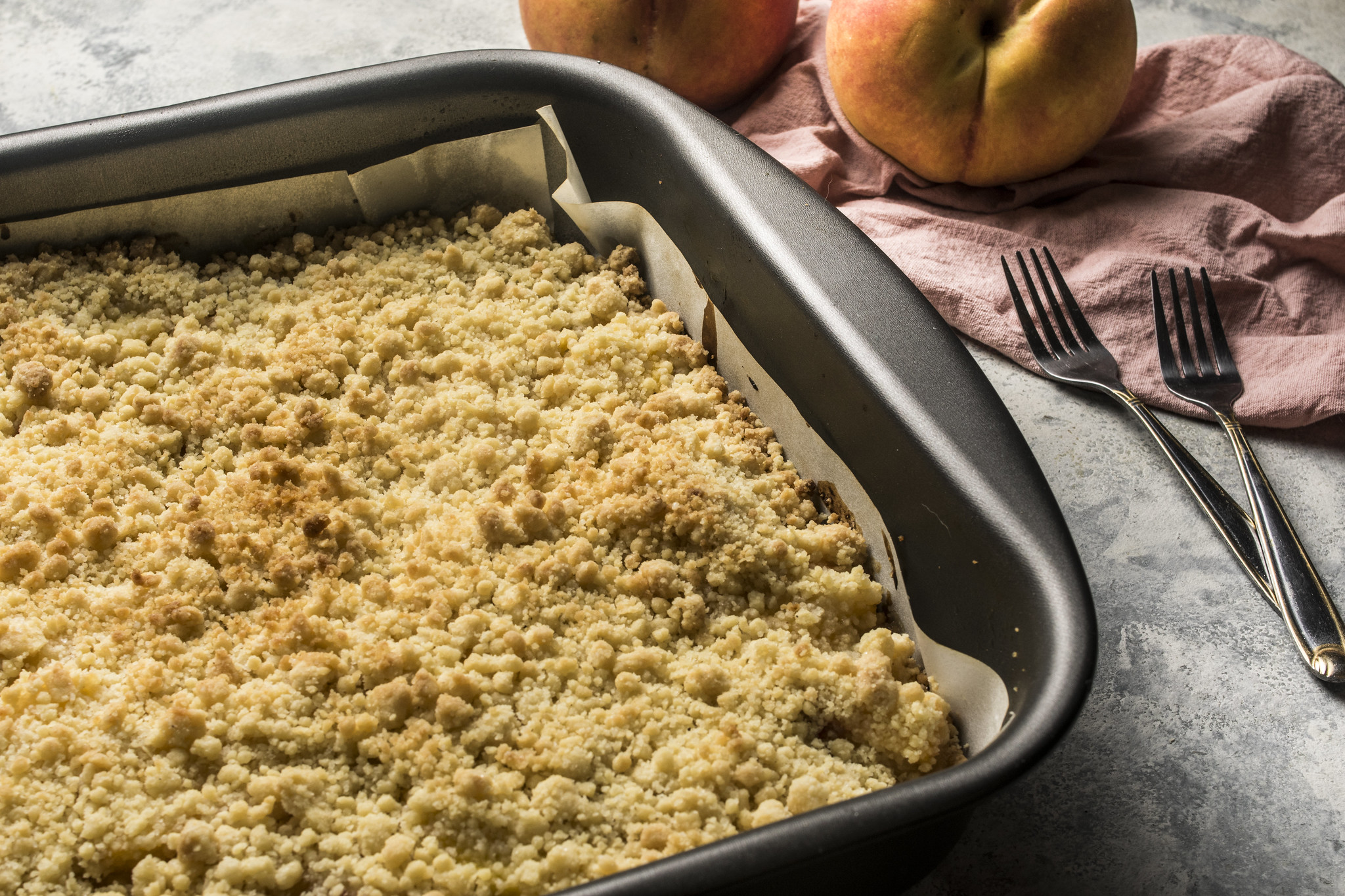 Freshly baked sbriciolata di pesche in a lined baking tin, with a golden crumb topping, peaches, pink cloth and vintage forks.
